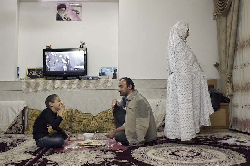 Noshabad, Iran, 16 novembre 2012. Una donna di religione musulmana sciita recita la quarta preghiera, quella della sera, mentre il resto della famiglia cena. Sullo sfondo la televisione accesa sulla diretta della cerimonia dell'Ashura a Karbala (Iraq).