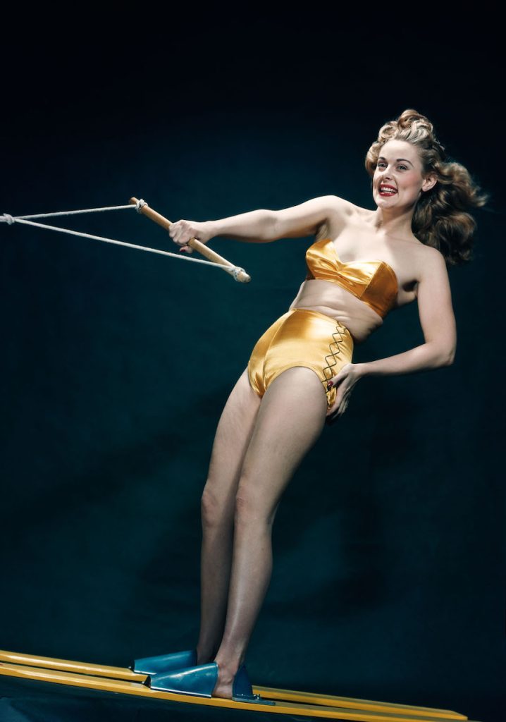 Smiling woman pinup wearing two piece gold bikini bathing suit posing riding water skis indoors Los Angeles