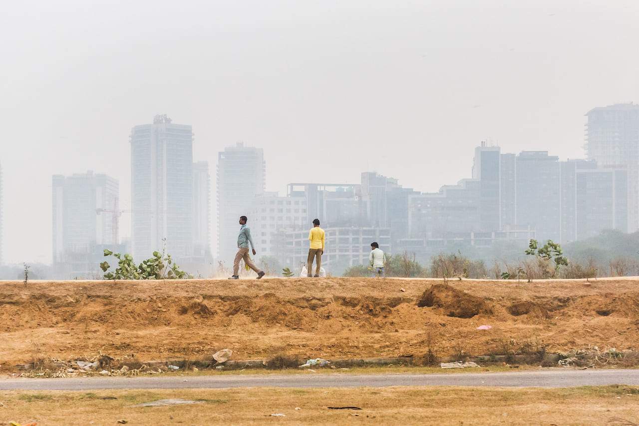 Alcuni uomini camminano in una strada periferica di Gurgaon, un centro tecnologico in rapida espansione alla periferia di Delhi, noto per avere uno dei peggiori indici di qualità dell'aria del pianeta. India, 2019 - Umidità 75%, Temperatura 21°C